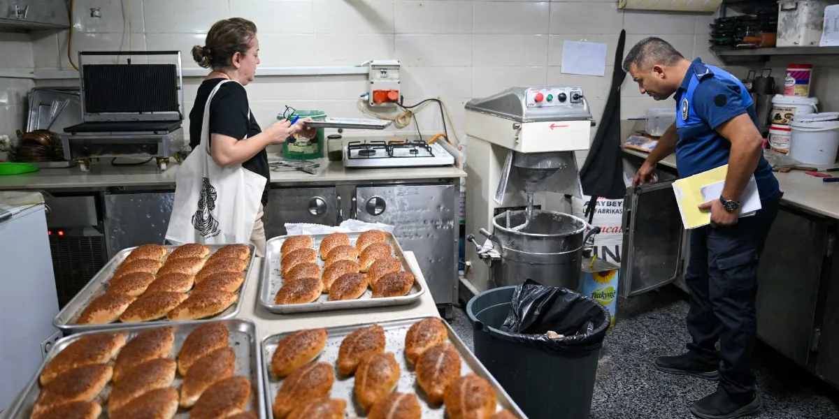 Bir fırın mutfağında, üzerinde taze pişmiş poğaçaların bulunduğu metal tepsiler tezgahın üzerinde dururken, mavi üniformalı bir zabıta memuru elindeki dosyalarla hamur yoğurma makinesini ve çevresini inceliyor. Yanında duran sivil kıyafetli bir kadın ise elinde boş bir tepsiyle denetim sürecine eşlik ediyor.