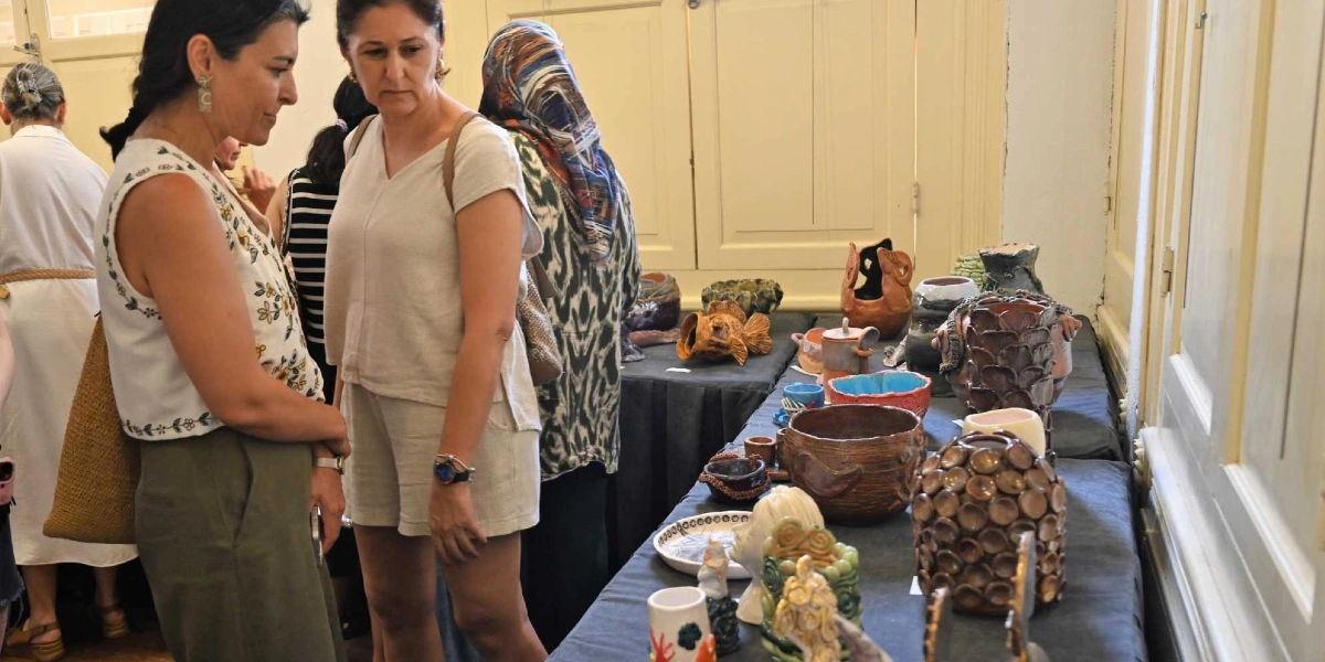 Bornova'daki bir sergide, masanın üzerinde sergilenen el yapımı seramik eserleri inceleyen kadınlar. Masanın üzerinde farklı formlarda, dokularda ve renklerde çömlekler, kaseler ve mitolojik figürleri andıran seramik objeler yer alıyor. Kursiyerlerin el emeği olan bu eserler, siyah örtü serili uzun bir masanın üzerinde, aydınlık bir iç mekanda sergileniyor.