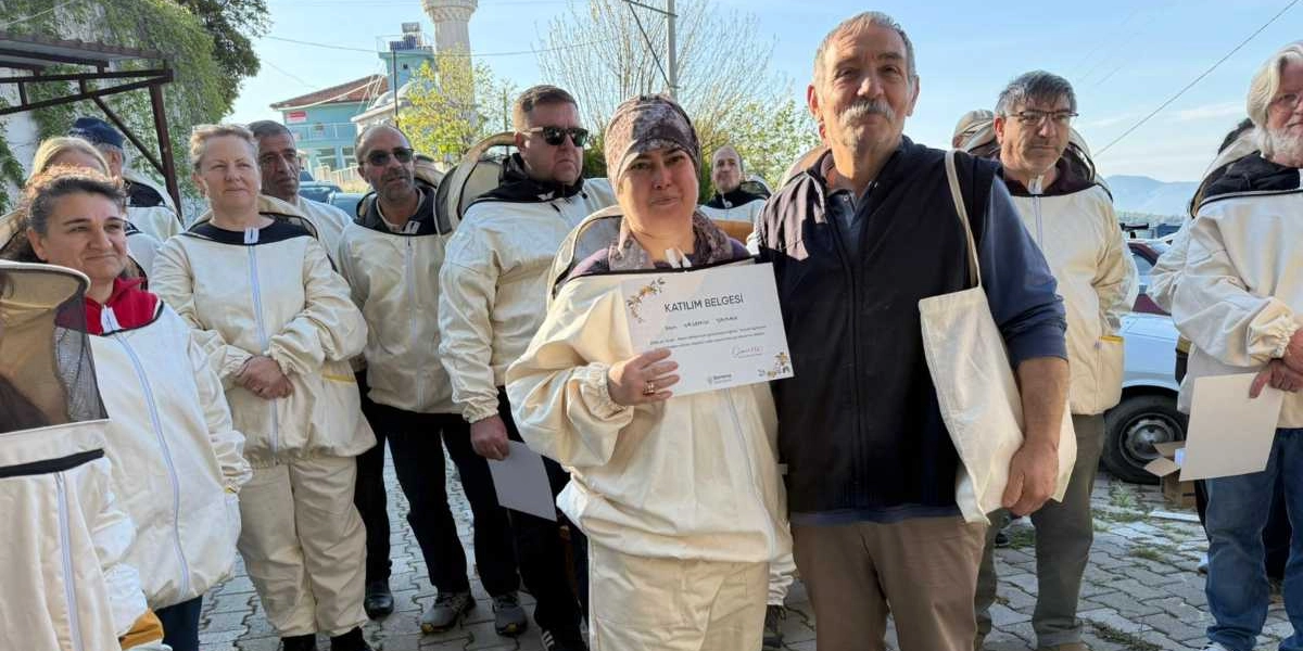 Arıcılık kıyafetleri giymiş bir grup insan, açık havada düzenlenen bir törende bir araya gelmiş. Grubun merkezinde, elinde 