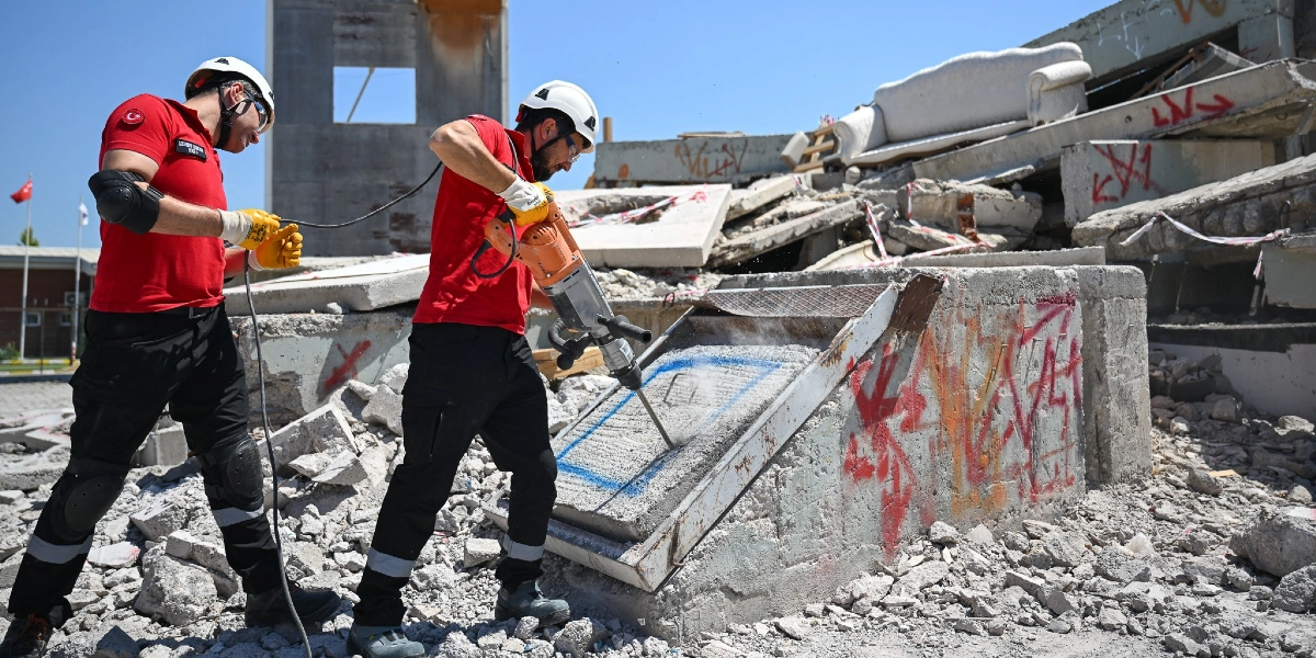 Kırmızı tişörtlü, beyaz baretli ve koruyucu ekipmanlı iki arama kurtarma görevlisi, yıkılmış bir binanın enkazında çalışıyor. Görevlilerden biri elindeki kırıcı delici matkapla beton bir bloğu kırmaya çalışırken, diğeri ona destek veriyor. Arka planda moloz yığınları ve üzerinde kırmızı sprey boya ile işaretler bulunan beton parçaları görülüyor.