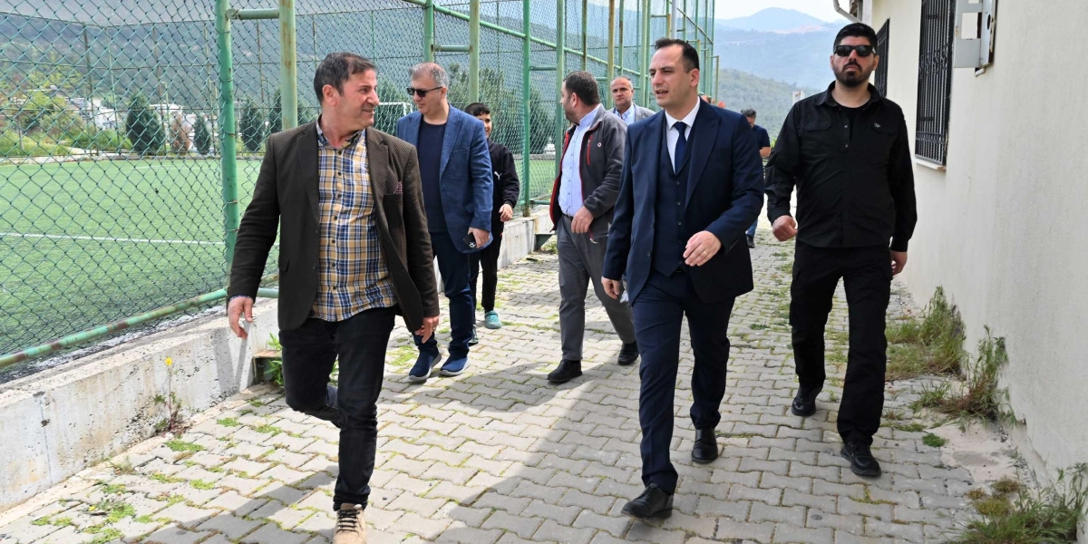 Lacivert takım elbiseli Başkan Eşki, yanında koruması ve beraberindeki heyetle birlikte, bir tarafında tel örgülü halı saha, diğer tarafında ise bir bina bulunan kaldırımda yürüyerek ilerliyor.