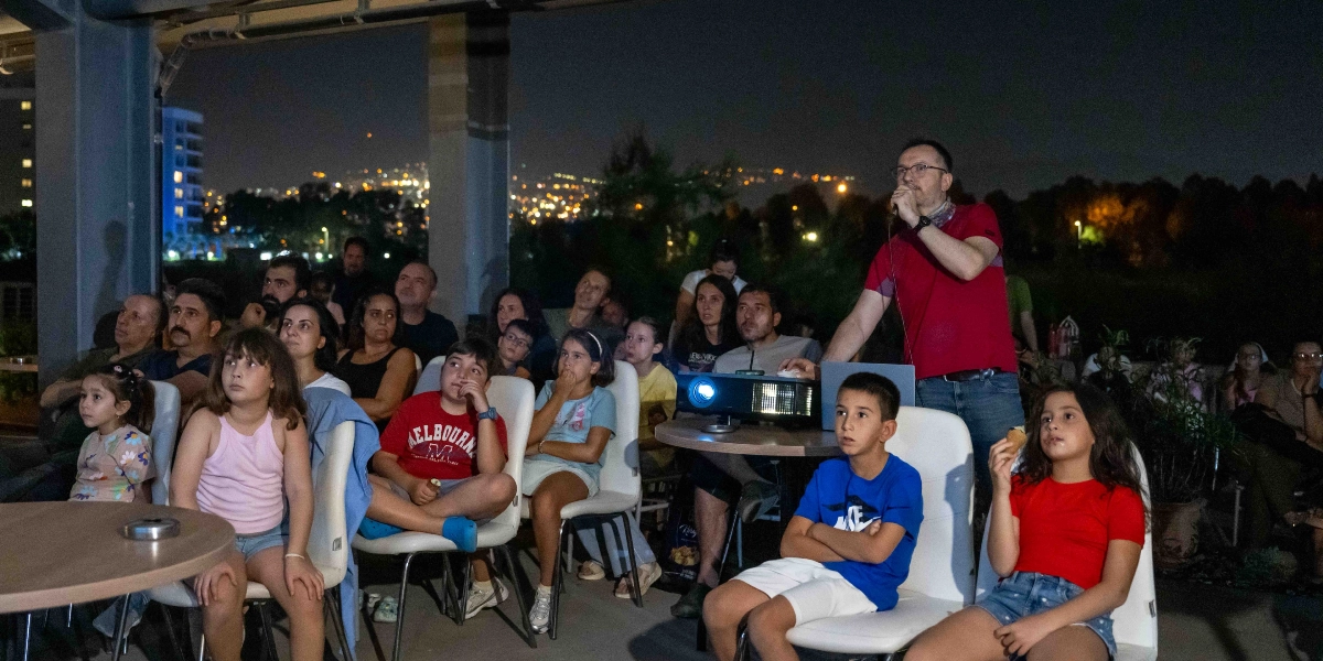 Gece vakti açık havada düzenlenen bir etkinlikte, kırmızı tişörtlü bir konuşmacı elinde mikrofonla izleyicilere hitap ediyor. Önündeki masada bir projeksiyon cihazı bulunuyor. İzleyici grubunda çocuklar ve yetişkinler yer alıyor; sandalyelerde oturmuş dikkatle konuşmacıyı dinliyorlar. Arka planda ise gece ışıklarıyla aydınlanan bir şehir manzarası görülüyor.