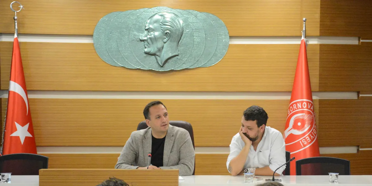 Ahşap panelli bir duvarın önünde, masada oturan iki erkek katılımcı. Arka planda duvarda Mustafa Kemal Atatürk'ün kabartma portresi, yanlarda ise Türk bayrağı ve üzerinde Bornova Belediyesi logosu bulunan kırmızı bir bayrak yer alıyor. Soldaki kişi gri ceketli ve konuşuyor, sağdaki kişi ise beyaz gömlekli ve elini çenesine dayamış şekilde dinliyor. Masanın üzerinde mikrofonlar ve su bardakları bulunuyor.