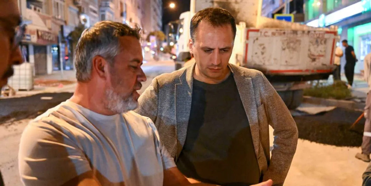 Gece vakti bir caddede, arka planda asfalt dökme çalışması yapan bir kamyonun önünde duran iki erkek, yapılan işi inceliyor. Öndeki beyaz tişörtlü ve kır saçlı adam bir şeyler anlatırken, yanındaki gri ceketli adam dikkatle onu dinliyor. Arka planda ise yol çalışması yapan diğer işçiler ve şehir ışıkları görülüyor.