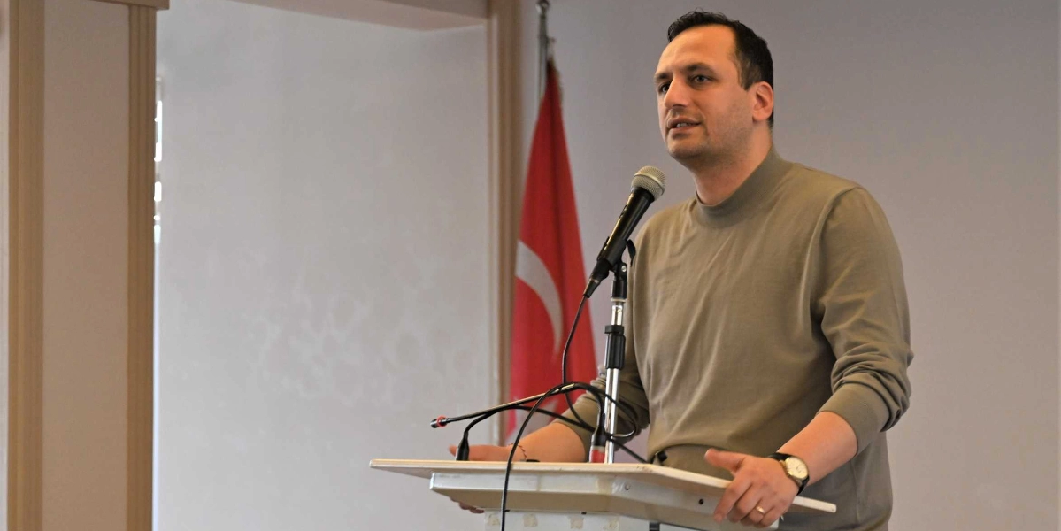 Haki renkli boğazlı kazak giymiş bir erkek, kürsüde mikrofon başında konuşma yapıyor. Arka planda Türk bayrağı yer alıyor.