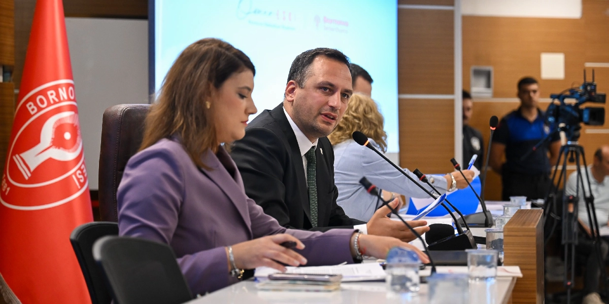 Bornova Belediye Başkanı Ömer Eşki, bir toplantı masasında yanında bir kadın yetkiliyle birlikte oturarak konuşma yapıyor. Masanın üzerinde mikrofonlar, belgeler ve su bardakları bulunuyor. Arka planda Bornova Belediyesi logosunun yer aldığı bir ekran ve sağ tarafta çekim yapan bir kameraman görülüyor.