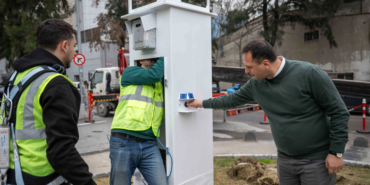 Dış mekanda, üzerinde güneş paneli benzeri küçük bir parça bulunan beyaz renkli dikey bir enerji ünitesinin kurulumunu yapan üç kişi. Yeşil kazaklı bir adam ünitenin üzerindeki bir düğmeye basarken, fosforlu iş yeleği giyen bir teknisyen ünitenin iç kısmıyla ilgileniyor; diğer tarafta ise emniyet kemeri takılı olan başka bir teknisyen çalışmaları izliyor.