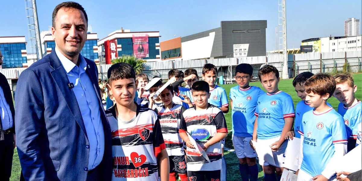 Lacivert ceketli ve açık mavi gömlekli bir yetkili, futbol sahasında ellerinde katılım belgeleri tutan çocuklardan oluşan bir grupla yan yana duruyor. Çocukların bir kısmı siyah-kırmızı çizgili, bir kısmı ise açık mavi renkli futbol formaları giyiyor. Arka planda binalar ve açık bir gökyüzü görülüyor.