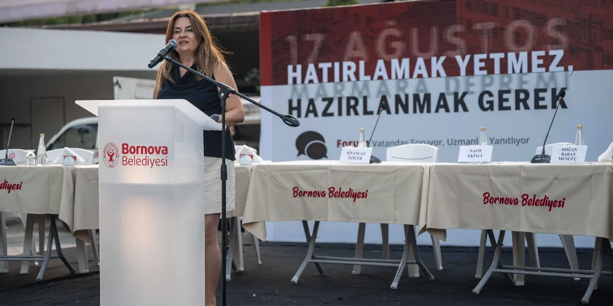 Bornova Belediyesi tarafından düzenlenen afet bilinci etkinliğinde, kürsüde konuşma yapan bir kadın yer alıyor. Arka planda üzerinde 17 Ağustos Hatırlamak Yetmez, Hazırlanmak Gerek yazılı büyük bir pano ve önünde üzerinde Bornova Belediyesi logolu örtülerin bulunduğu boş bir panel masası bulunuyor. Masanın üzerinde Sinancan Öziçer, Nazif Ekinci ve Doğan Baran Mengüç isimlerinin yazılı olduğu isimlikler yer alıyor.