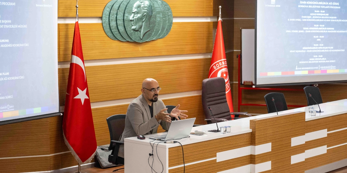 Bornova Belediyesi'nde düzenlenen İzmir Sürdürülebilirlik Ağı toplantısında, kürsüde oturan bir konuşmacı dizüstü bilgisayarı eşliğinde sunum yapıyor. Arka planda duvarda Atatürk rölyefi, iki yanında Türk bayrağı ve Bornova Belediyesi bayrağı yer alıyor. Yanlarda ise toplantı programının yansıtıldığı iki büyük projeksiyon ekranı bulunuyor.