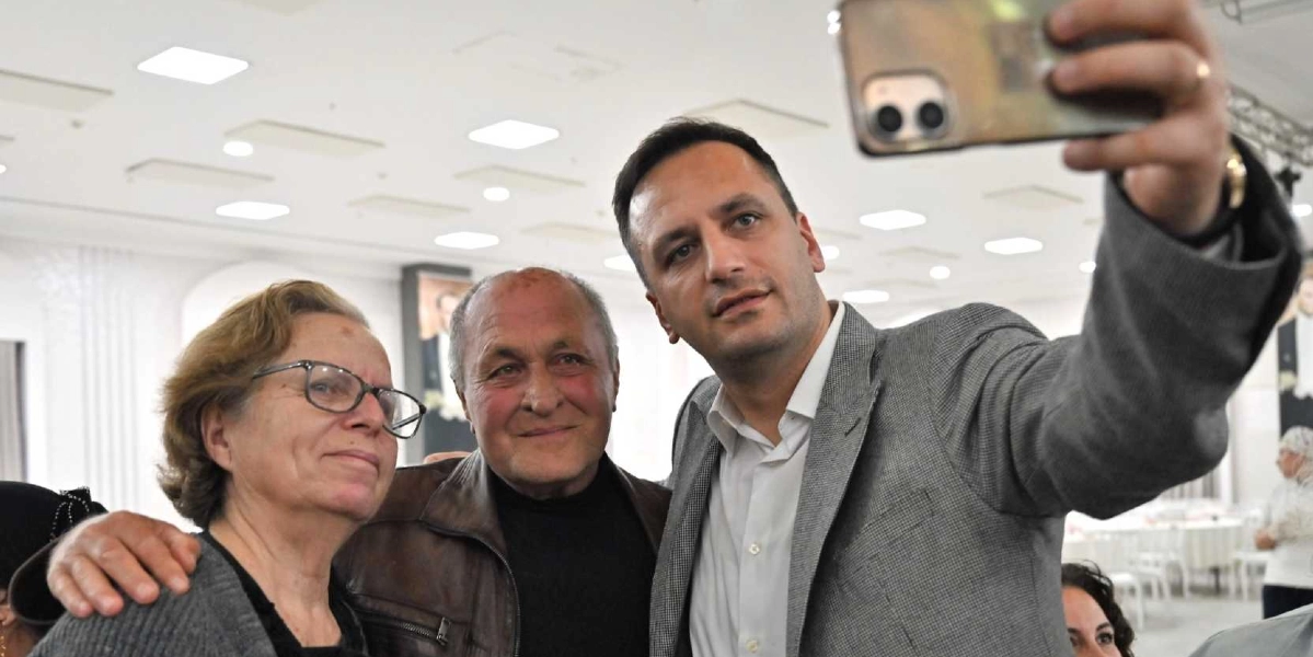 Gri ceketli bir erkek, yanında duran orta yaşlı bir kadın ve bir erkekle birlikte gülümseyerek selfie çekiyor. Arka planda aydınlık, geniş bir salon ve yemek masaları görülüyor.