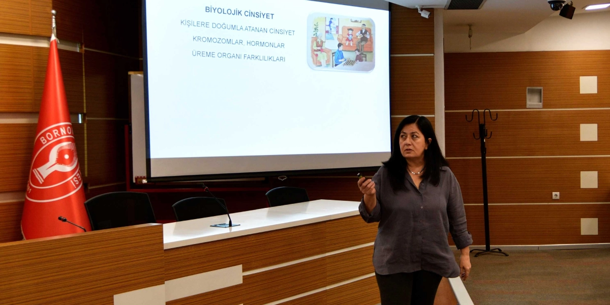 Bornova Belediyesi'ne ait bir toplantı salonunda, elinde sunum kumandasıyla konuşma yapan orta yaşlı bir kadın yer alıyor. Kadının arkasındaki projeksiyon perdesinde 