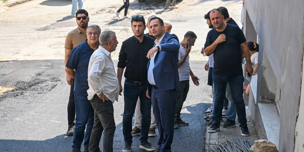 Mavi takım elbiseli bir belediye başkanı, yeni asfaltlanmış bir sokakta yanındaki heyetle birlikte çalışmaları inceliyor ve eliyle bir noktayı işaret ederek açıklamalarda bulunuyor. Çevresinde farklı kıyafetler giymiş erkeklerden oluşan bir grup, dikkatle başkanı dinliyor.