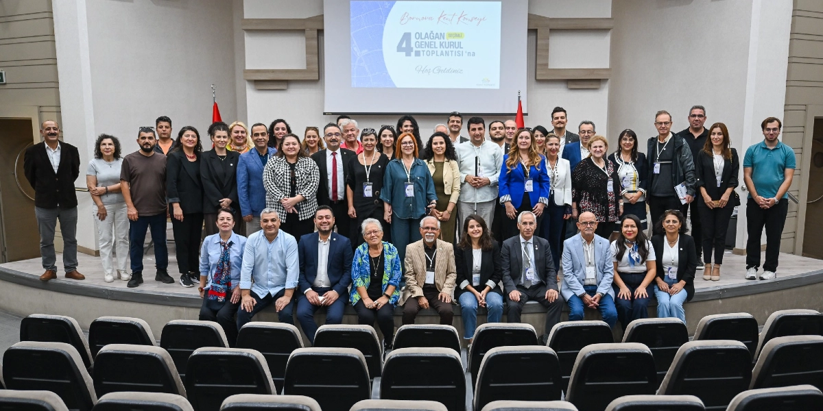 Bornova Kent Konseyi 4. Olağan Seçimli Genel Kurul Toplantısı için bir araya gelen, aralarında farklı yaş ve meslek gruplarından kadın ve erkeklerin bulunduğu kalabalık bir grup, sahne üzerinde yan yana dizilerek poz veriyor. Arka planda üzerinde 
