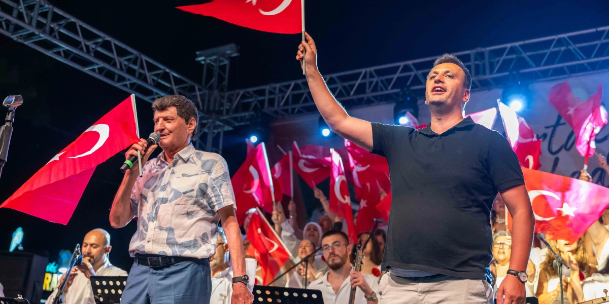 Gece vakti açık hava sahnesinde, ellerinde Türk bayrakları tutan bir grup müzisyen ve şarkıcı performans sergiliyor. Ön planda, sol tarafta kısa kollu desenli bir gömlek giyen bir kişi mikrofonla şarkı söylerken, sağ tarafta siyah polo yaka tişörtlü bir kişi elini havaya kaldırarak coşkuyla eşlik ediyor. Arka planda ise koro üyeleri ve enstrüman çalan müzisyenler yer alıyor; sahne ışıkları ve sallanan bayraklar şenlikli bir atmosfer oluşturuyor.