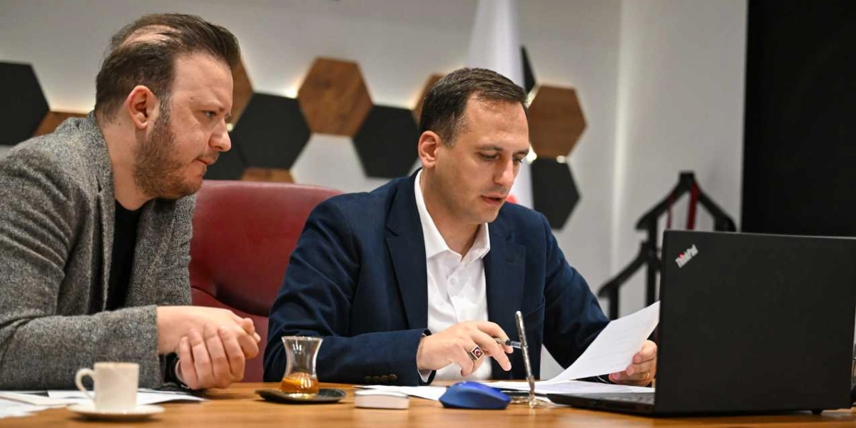 Ofis ortamında bir masada oturan iki erkek, önlerindeki dizüstü bilgisayara ve belgelere bakarak enerji projeleri üzerine çalışıyor. Lacivert ceketli ve beyaz gömlekli kişi elinde bir belge tutarken, yanındaki gri ceketli kişi onu dikkatle dinliyor. Masanın üzerinde bir bardak çay, bir fincan kahve ve teknik ekipmanlar bulunuyor. Arka planda ise altıgen desenli duvar dekorasyonu yer alıyor.