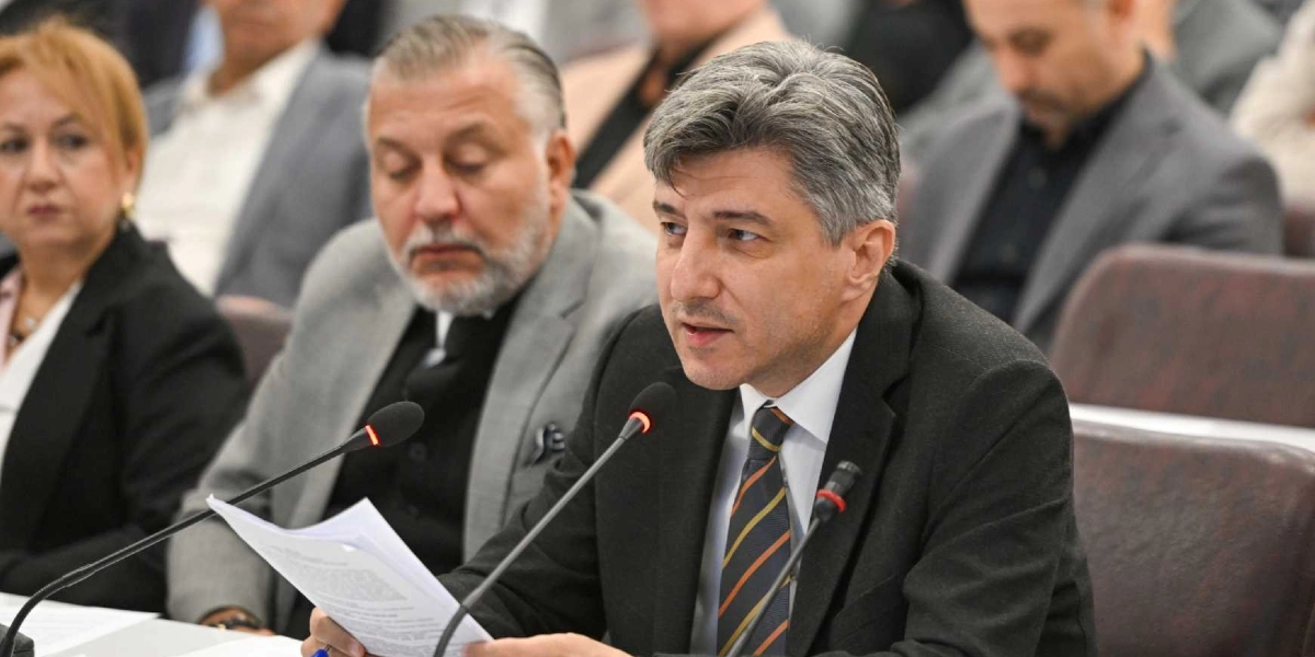 Bornova Belediyesi meclis toplantısında, siyah takım elbiseli ve çizgili kravatlı bir yetkili mikrofon başında elindeki belgeleri okuyarak konuşma yapıyor. Yanında gri ceketli, sakallı bir beyefendi ve sol tarafta sarı saçlı bir kadın yetkili oturuyor. Arka planda toplantıya katılan diğer meclis üyeleri yer alıyor.