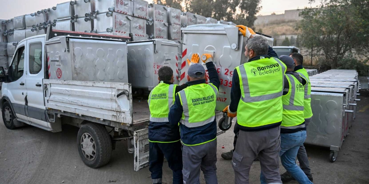 Bornova Belediyesi Temizlik İşleri Müdürlüğü personeli, üzerinde belediye logosu bulunan fosforlu sarı yelekleriyle, kamyondan yeni metal çöp konteynerlerini indiriyor. Arka planda üst üste dizilmiş çok sayıda boş konteyner bulunuyor.