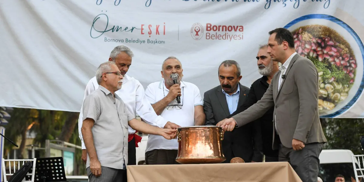 Bornova Belediye Başkanı Ömer Eşki ve beraberindeki beş kişi, üzerinde Bornova Belediyesi logosu ve aşure görseli bulunan bir pankartın önünde, masanın üzerinde duran büyük bakır bir kazanın etrafında toplanmışlar. Gruptan biri mikrofonla konuşurken, diğerleri ellerini kazanın üzerine koyarak sembolik bir aşure karıştırma etkinliği gerçekleştiriyor.