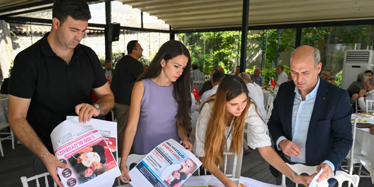 Dramalılar Köşkü'ndeki bir toplantıda, siyah tişörtlü bir erkek, lila elbiseli bir kadın, beyaz dantelli bluzlu bir kadın ve lacivert ceketli bir erkek, ellerinde SMA hastası Abbas Miran için yardım çağrısı içeren broşürleri inceliyor. Arka planda masalarda oturan diğer katılımcılar ve yeşilliklerle çevrili bir bahçe ortamı görülüyor.