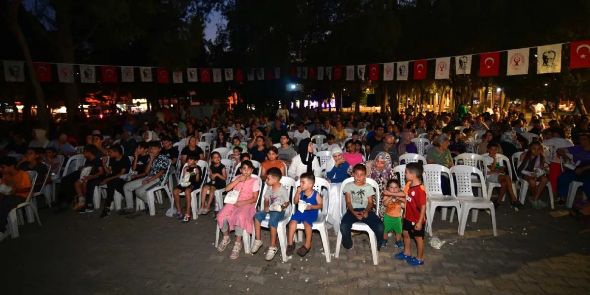 Açık havada, akşam saatlerinde düzenlenen bir film gösteriminde beyaz plastik sandalyelerde oturan kalabalık bir izleyici grubu. Park ortamındaki alanda çocuklar ve yetişkinler yan yana oturarak ekranı izliyor. Alanın üzerinde, Türk bayrakları ve Atatürk portrelerinin yer aldığı flamalar dizili bir şekilde asılı duruyor.