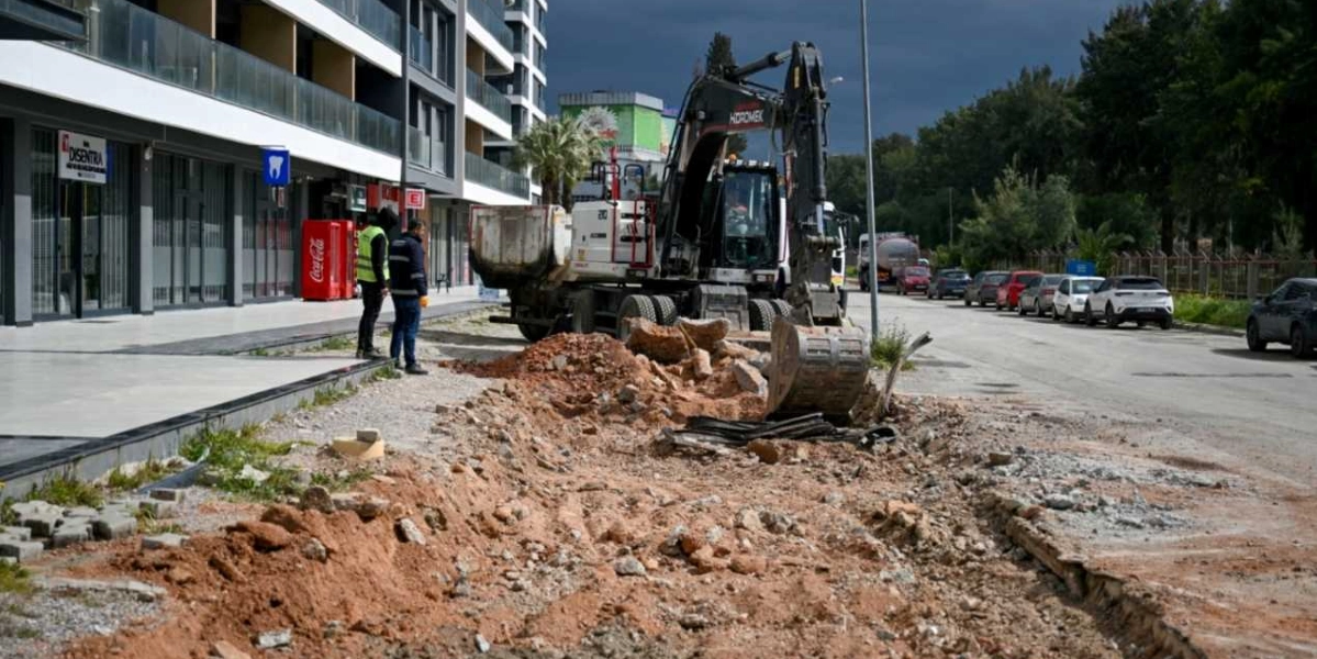 Bornova'da bir cadde kenarında devam eden yol yapım çalışması. Sarı yelekli iki görevli, kazı yapılan alanın başında durarak çalışmaları izliyor. Büyük bir iş makinesi, kaldırım kenarındaki toprak ve moloz yığınlarını temizliyor. Yolun sol tarafında modern bir apartman, sağ tarafında ise park halindeki araçlar görülüyor.