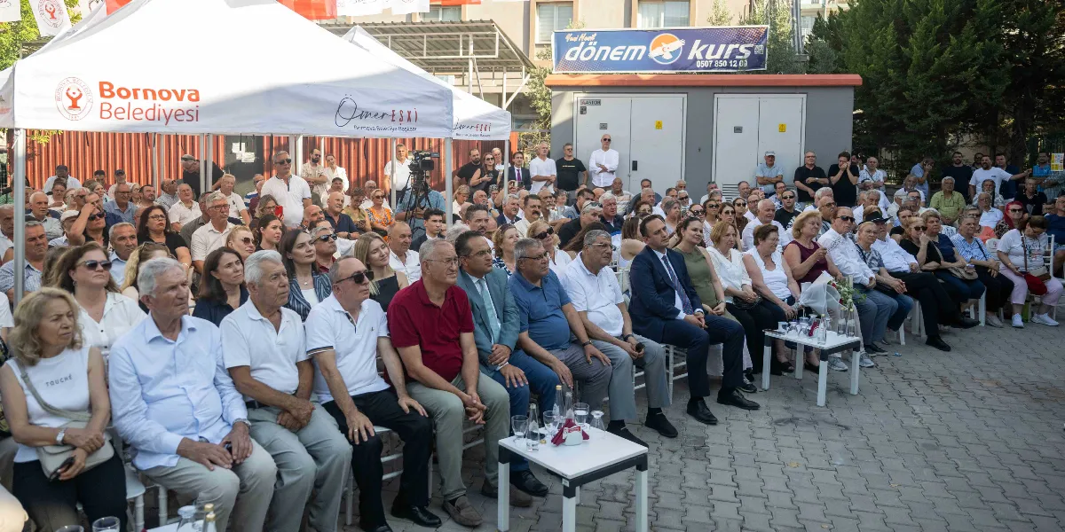 Açık havada, Bornova Belediyesi logolu beyaz bir çadırın altında ve çevresinde toplanmış, Dr. Şaban Acarbay'ın isminin verileceği yüzme havuzu için düzenlenen töreni izleyen kalabalık bir grup insan. Katılımcılar sandalyelerde oturmuş, önlerinde küçük masalar ve su şişeleri bulunan bir alanda etkinliği takip ediyor. Arka planda bir binanın duvarında 