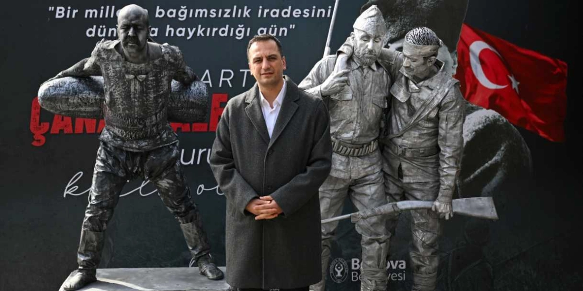 Gri palto giymiş bir erkek, arkasında Çanakkale Zaferi'ni anımsatan gümüş renkli asker heykellerinin ve üzerinde 