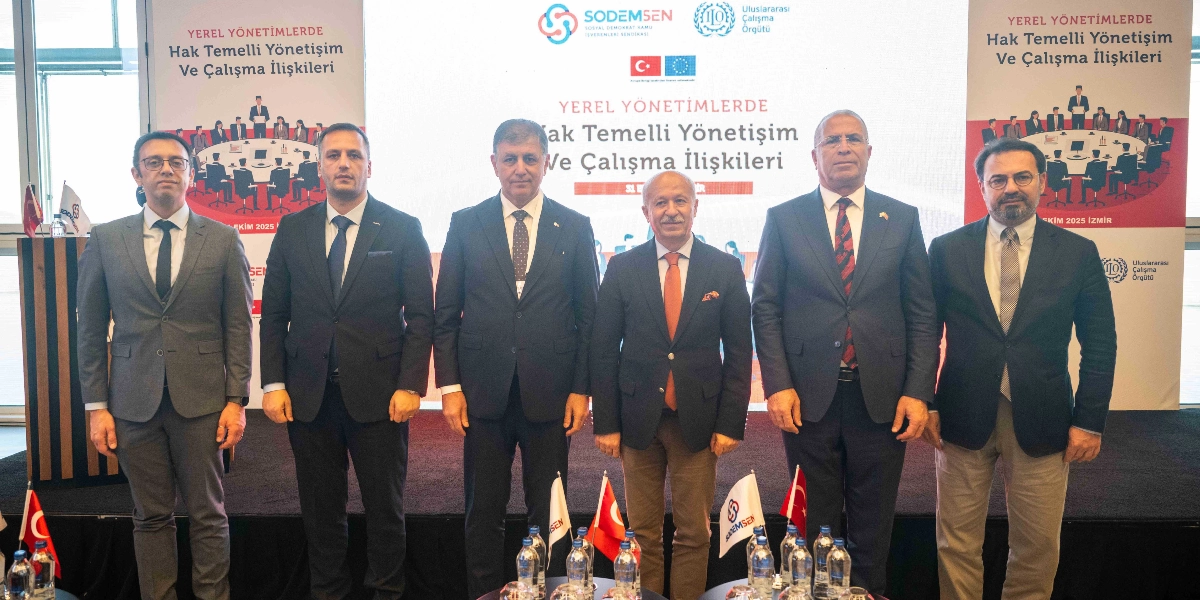 SODEMSEN tarafından düzenlenen Yerel Yönetimlerde Hak Temelli Yönetişim ve Çalışma İlişkileri çalıştayında, sahnedeki dev ekranın önünde yan yana duran altı erkek katılımcı. Katılımcıların çoğu koyu renk takım elbise ve kravat giyerken, ortadaki kişi turuncu kravat ve açık renk pantolonla yer alıyor. Arka planda SODEMSEN ve Uluslararası Çalışma Örgütü logolarının bulunduğu, üzerinde etkinliğin adının yazılı olduğu büyük bir ekran ve iki yanda aynı başlığı taşıyan dikey afişler yer alıyor. Ön plandaki masada küçük Türk bayrakları ve SODEMSEN logolu bayraklar ile su şişeleri bulunuyor.