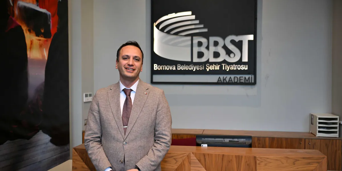 Bej renkli ceket ve kravat giymiş, gülümseyen bir erkek, arkasında Bornova Belediyesi Şehir Tiyatrosu (BBST) Akademi yazılı tabelanın bulunduğu bir resepsiyon bankosunun önünde duruyor.