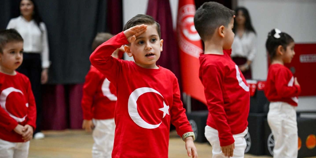 Üzerlerinde Türk bayrağı baskılı kırmızı tişörtler ve beyaz pantolonlar bulunan anaokulu çağındaki çocuklar, bir gösteri sırasında asker selamı veriyor. Arka planda Türk bayrakları ve yetişkinler yer alıyor.