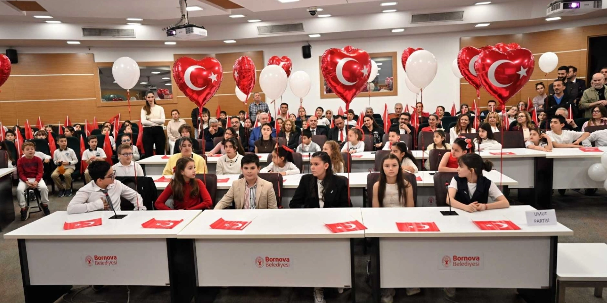 Bornova Belediyesi'nin bir toplantı salonunda düzenlenen 23 Nisan Ulusal Egemenlik ve Çocuk Bayramı etkinliğinde, sıralarda oturan çocuklar ve arkalarında onları izleyen yetişkinler yer alıyor. Çocukların oturduğu masaların üzerinde Türk bayrakları ve 