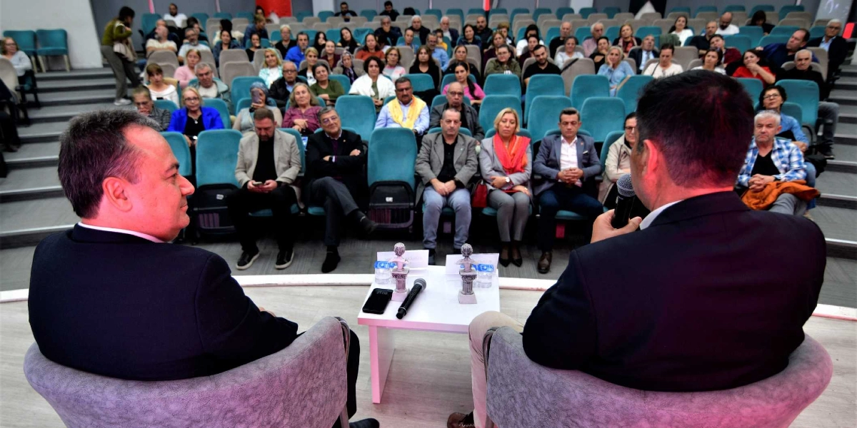 Konferans salonunda sahnedeki iki konuşmacı, karşılarında oturan kalabalık bir dinleyici grubuna hitap ediyor. Ön planda, arkaları dönük şekilde gri koltuklarda oturan iki erkek konuşmacı bulunuyor; sağdaki konuşmacı elinde mikrofon tutuyor. Aralarındaki küçük beyaz masanın üzerinde iki adet küçük heykelcik, su şişeleri ve bir mikrofon yer alıyor. Arka planda, kademeli olarak yükselen turkuaz renkli koltuklarda oturan çok sayıda katılımcı, sahnedeki konuşmacıları dikkatle dinliyor.