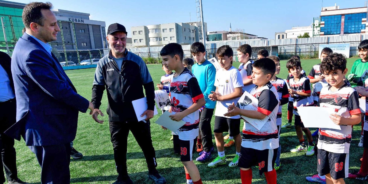 Açık havada, suni çim kaplı bir futbol sahasında, takım formaları giymiş bir grup çocuk futbolcuya yetkililer tarafından üzerinde Atatürk fotoğrafı bulunan başarı belgeleri takdim ediliyor. Lacivert takım elbiseli bir beyefendi ve siyah eşofmanlı bir antrenör, çocuklara belgelerini verirken gülümsüyor. Çocuklar ellerinde belgelerle yan yana dizilmiş, neşeli bir şekilde bekliyor. Arka planda binalar ve çitlerle çevrili bir spor alanı görülüyor.
