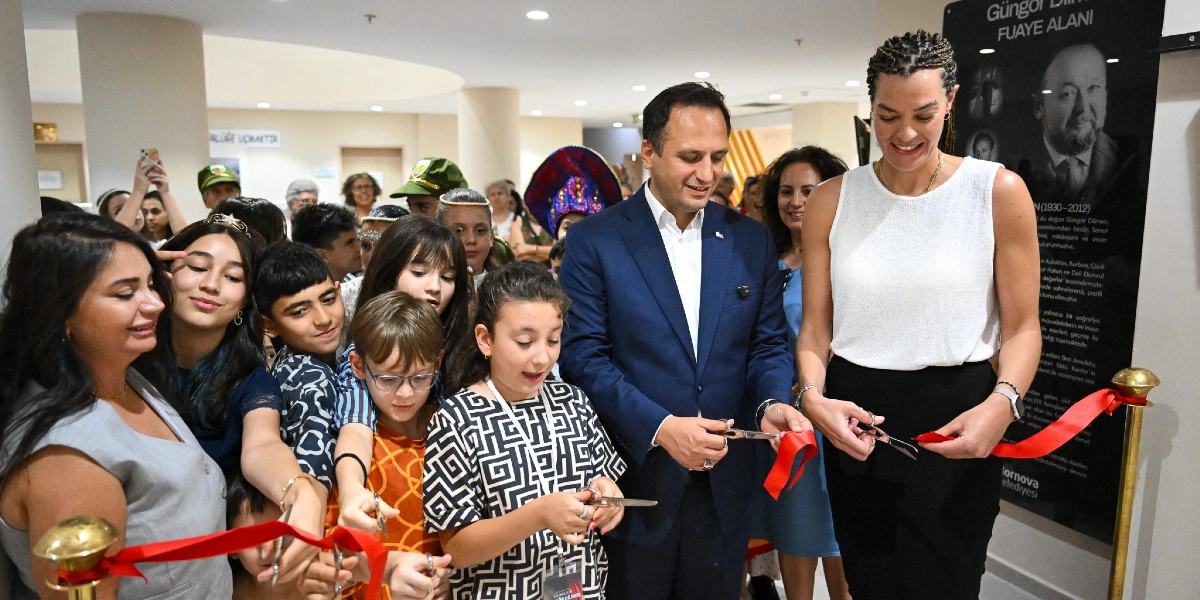 Bornova Belediye Başkanı Mustafa İduğ ve yanında bulunan bir kadın, bir grup çocukla birlikte Güngör Dilmen Fuaye Alanı'nın açılış kurdelesini kesiyor. Modern bir binanın iç mekanında gerçekleşen törende, çocuklar heyecanla kurdeleyi tutarken, arka planda izleyiciler yer alıyor. Sağ taraftaki duvarda Güngör Dilmen'in portresinin ve biyografisinin yer aldığı siyah bir bilgilendirme panosu bulunuyor.