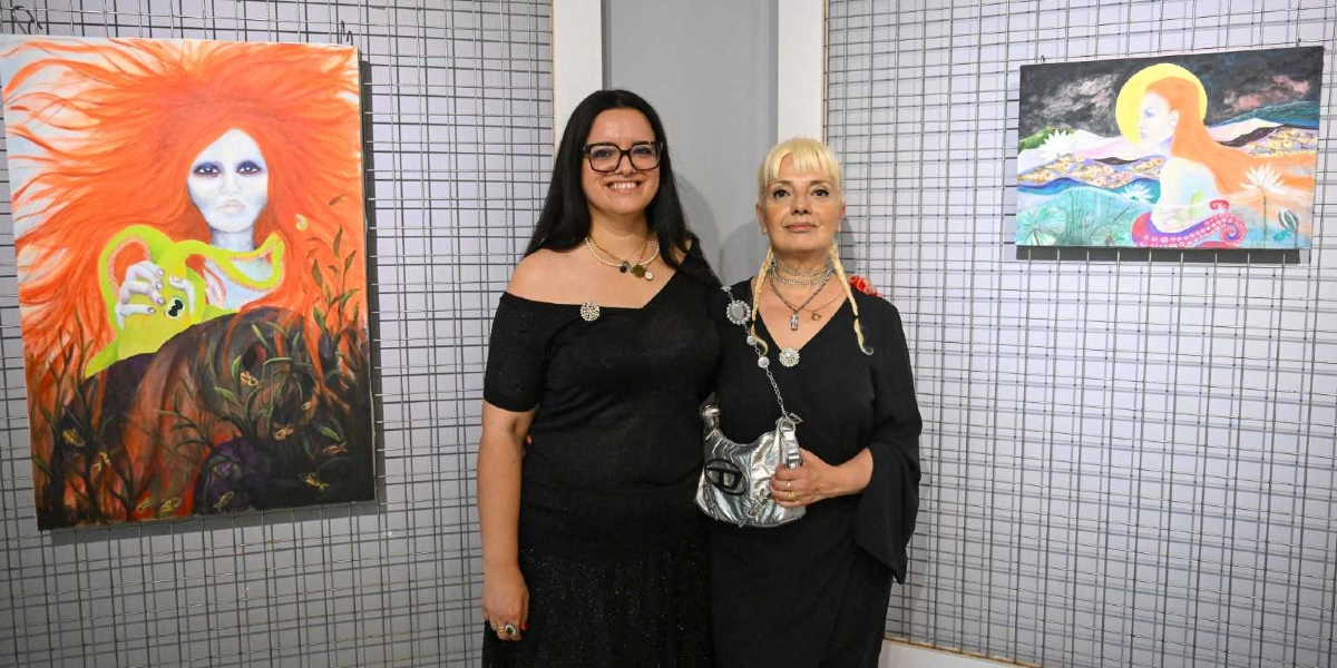 Bir sanat galerisinde, ızgara panelli duvarın önünde yan yana duran iki kadın. Soldaki kadın uzun siyah saçlı, gözlüklü ve siyah bir bluz giyiyor; yanında turuncu saçlı, fantastik bir figürün olduğu büyük bir tablo yer alıyor. Sağdaki kadın sarı saçlı, siyah elbiseli ve omzunda gümüş renkli bir çanta taşıyor; onun yanında ise gökyüzünde sarı bir küre ve figürlerin yer aldığı daha küçük bir tablo bulunuyor.