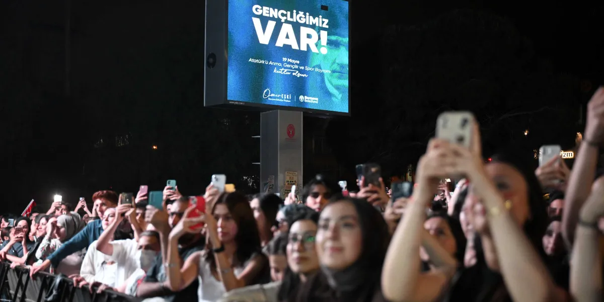 Gece vakti bir konser veya etkinlik alanında, ellerinde telefonlarıyla sahneyi kaydeden kalabalık bir genç grubu görülüyor. Arka planda yüksek bir dijital ekranda, üzerinde Atatürk silüeti bulunan ve 