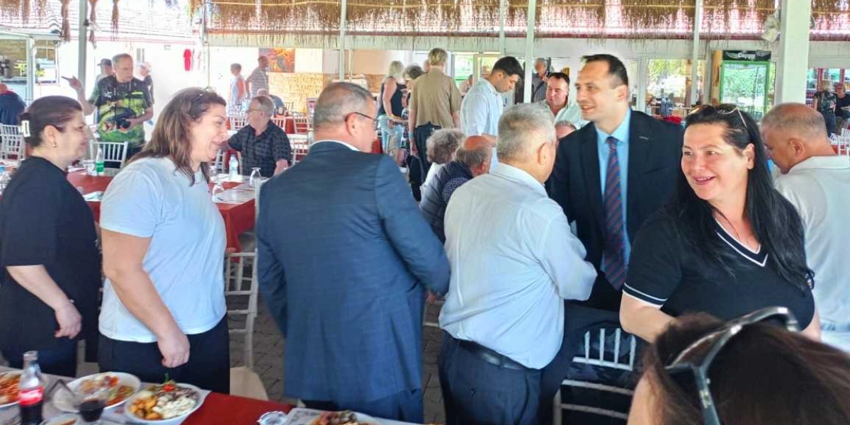 Kapalı bir mekanda, masaların bulunduğu bir ortamda bir araya gelmiş, aralarında Bornova Muhtarlar Akademisi katılımcılarının da olduğu tahmin edilen bir grup insan. Ön planda beyaz tişörtlü bir kadın, lacivert takım elbiseli bir erkek, açık renk gömlekli başka bir erkek ve siyah bluzlu bir kadın birbirleriyle sohbet ediyor. Arka planda ise ayakta duran veya masalarda oturan diğer katılımcılar ve yemek tabakları görülüyor.