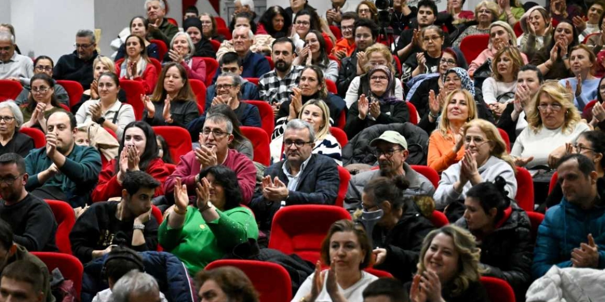 Kapalı bir salonda, kırmızı koltuklarda oturan ve büyük bir ilgiyle alkışlayan kalabalık bir izleyici grubu. Farklı yaş gruplarından kadın ve erkeklerin yer aldığı izleyiciler, sahnedeki bir performansı veya konuşmayı dikkatle takip ederek desteklerini alkışlarla gösteriyor.