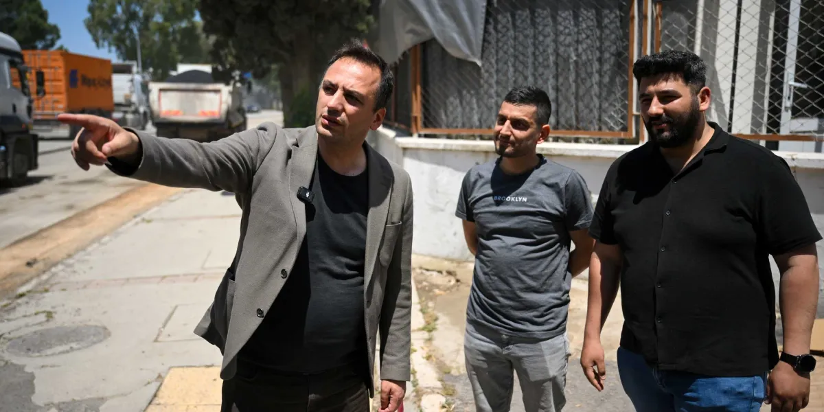 Gri ceketli bir adam, yanında duran iki kişiye eliyle caddenin ilerisini işaret ederek bir şeyler anlatıyor. Arka planda Pınar Caddesi'ndeki yol çalışmaları ve park halindeki ağır vasıta araçları görülüyor.