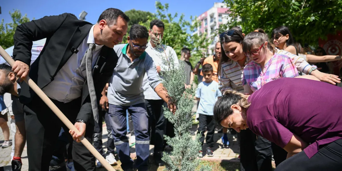 Açık havada, güneşli bir günde düzenlenen fidan dikme etkinliğinde, takım elbiseli bir erkek elindeki kürekle toprağı kazarken, yanındaki katılımcılar ve özel gereksinimli bireyler fidanın dikilmesine yardımcı oluyor. Çevredeki insanlar dikilen küçük çam ağacını ilgiyle izliyor ve destek veriyor.
