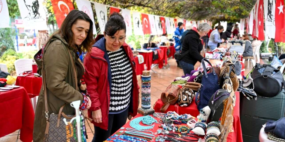 Açık havada kurulan bir hediyelik eşya pazarında, kırmızı örtülü tezgahlarda sergilenen el yapımı takıları ve çantaları inceleyen iki kadın. Arka planda, üzerinde Türk bayrakları ve Atatürk görselleri bulunan flamaların asılı olduğu bir yürüyüş yolu ve alışveriş yapan diğer insanlar görülüyor.