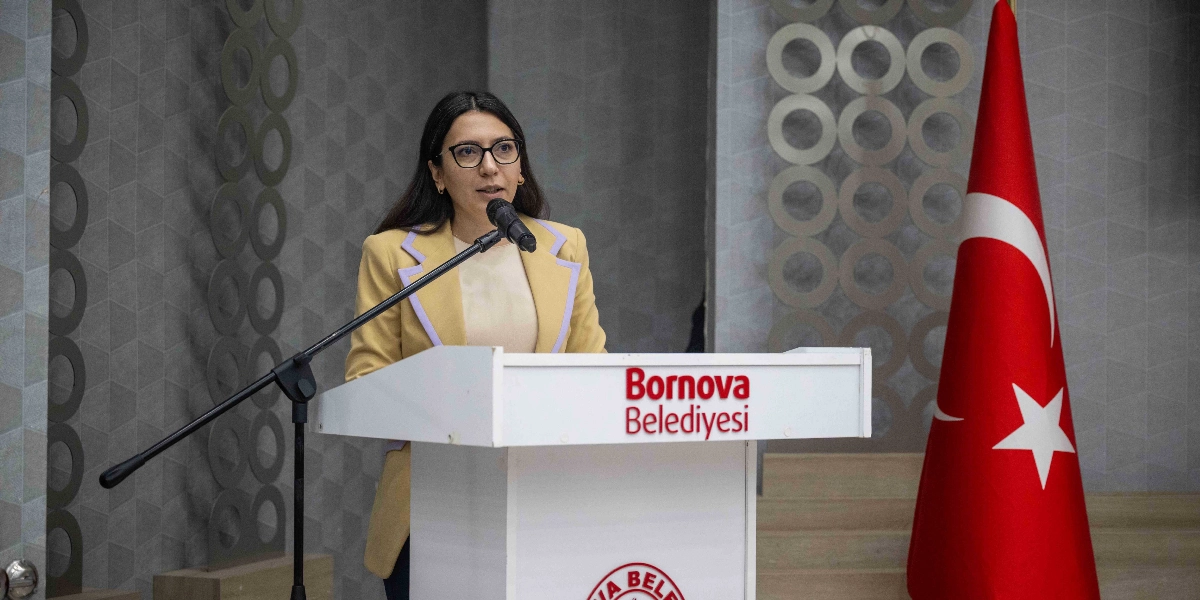 Gözlüklü, uzun koyu renk saçlı bir kadın, üzerinde Bornova Belediyesi yazan bir kürsüde mikrofon başında konuşma yapıyor. Kadın, açık sarı renkli, yaka kısımları farklı tonda bir ceket giyiyor. Kürsünün hemen sağ tarafında ise bir Türk bayrağı yer alıyor.