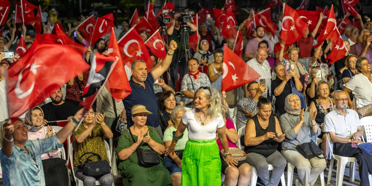 Açık havada düzenlenen bir etkinlikte, ellerinde Türk bayrakları tutan ve coşkuyla alkışlayan kalabalık bir grup insan. İzleyiciler arasında beyaz tişörtlü ve yeşil etekli bir kadın ayakta durarak neşeyle dans ederken, arkasında oturan ve ayakta duran diğer katılımcılar bayrak sallayarak ona eşlik ediyor. Arka planda profesyonel kamera ekipmanları ve etkinliği cep telefonlarıyla kaydeden izleyiciler görülüyor.