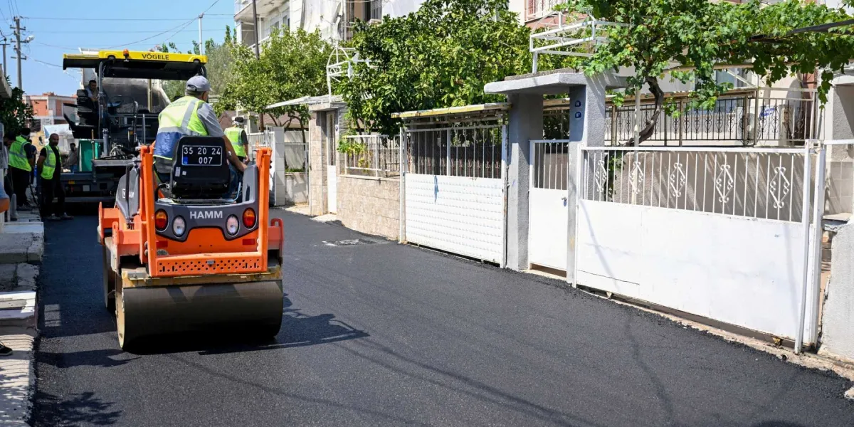 Belediye ekipleri tarafından yerleşim yerindeki bir sokakta gerçekleştirilen asfalt serme ve sıkıştırma çalışması. Turuncu renkli Hamm marka bir silindir, yeni dökülmüş asfalt üzerinde ilerlerken, arkasında Vögele marka bir asfalt serici iş makinesi ve yelekli görevliler çalışmaya devam ediyor.