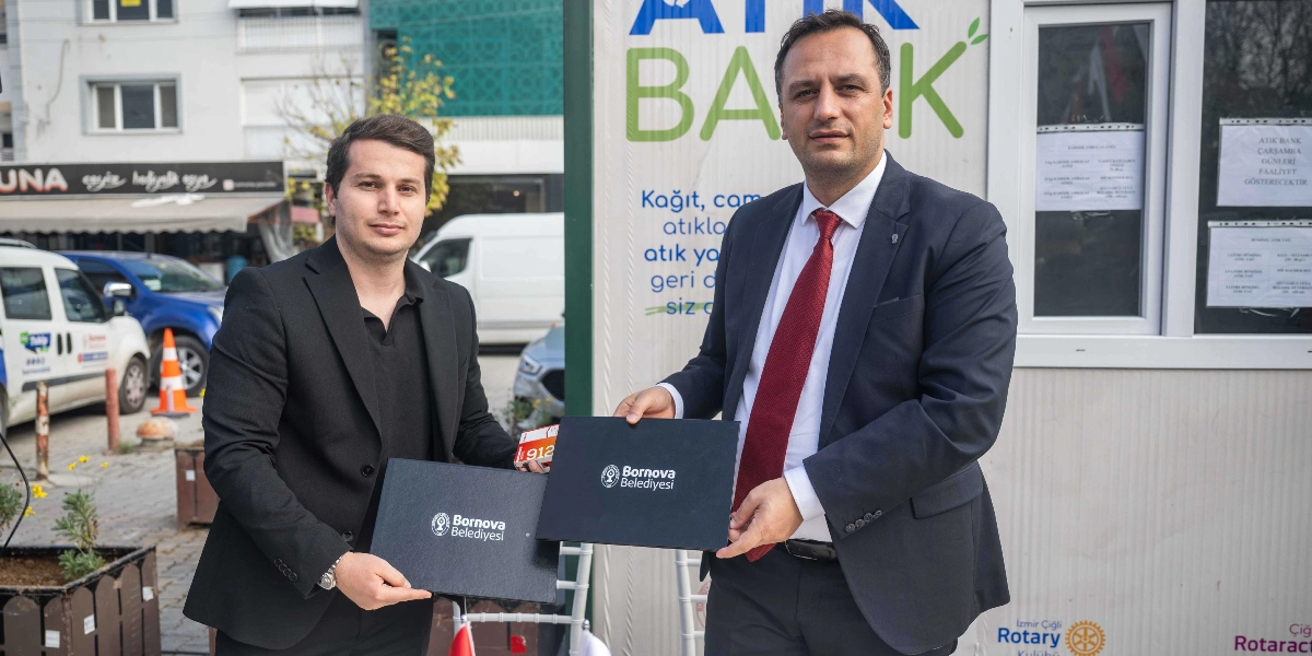 Bornova Belediyesi tarafından hayata geçirilen Atık Bank projesinin açılışında, üzerinde Bornova Belediyesi logosu bulunan siyah dosyaları birbirine takdim eden iki erkek yer alıyor. Soldaki kişi siyah ceket ve siyah tişört giymiş, sağdaki kişi ise lacivert takım elbise ve kırmızı kravat takmış durumda. Arkalarında 