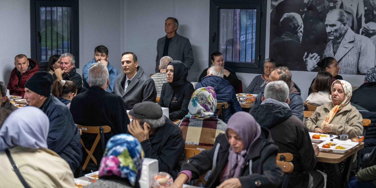 Kapalı bir mekanda, masalarda oturan ve önlerinde yemek tepsileri bulunan kalabalık bir grup insan iftar yemeği için bir araya gelmiş. Aralarında başörtülü kadınların ve yaşlıların da bulunduğu katılımcılar, masalarda yan yana oturarak yemeklerini yiyor. Arka planda duvarda Mustafa Kemal Atatürk'ün siyah beyaz bir fotoğrafı asılı duruyor.