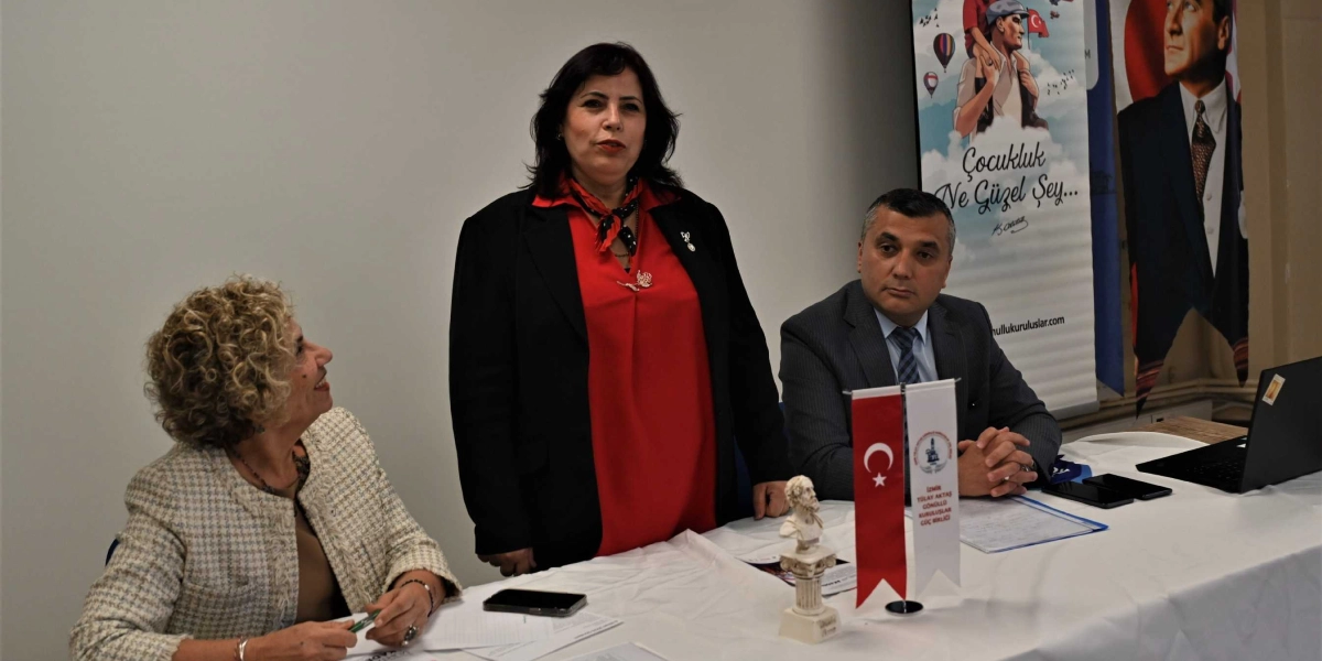 Bir toplantı masasında oturan üç kişi; solda açık renkli ceketli bir kadın, ortada kırmızı bluz ve siyah ceket giymiş ayakta duran bir kadın ve sağda takım elbiseli bir erkek yer alıyor. Masanın üzerinde küçük bir Türk bayrağı, İzmir Tülay Aktaş Gönüllü Kuruluşlar Güç Birliği yazılı bir flama ve küçük bir büst bulunuyor. Arka planda üzerinde 