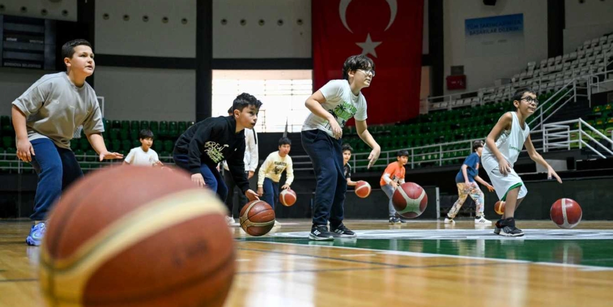 Kapalı bir spor salonunda basketbol antrenmanı yapan bir grup çocuk, ellerinde toplarla sahada duruyor. Ön planda odak dışı büyük bir basketbol topu yer alırken, arka planda antrenörleri eşliğinde top sürme çalışması yapan çocuklar ve duvarda asılı büyük bir Türk bayrağı görülüyor.