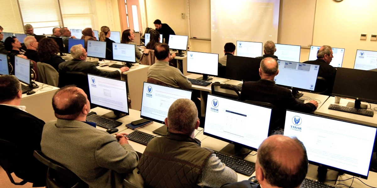 Yaşar Üniversitesi bilgisayar laboratuvarında düzenlenen eğitimde, sıralarda oturan muhtarlar bilgisayar ekranlarına bakarak dersi takip ediyor. Sınıfın ön tarafında bir eğitmen ayakta dururken, projeksiyon cihazından yansıtılan ekran görüntüsü ve katılımcıların önündeki bilgisayar monitörlerinde Yaşar Üniversitesi logosu ve eğitim içerikleri yer alıyor.